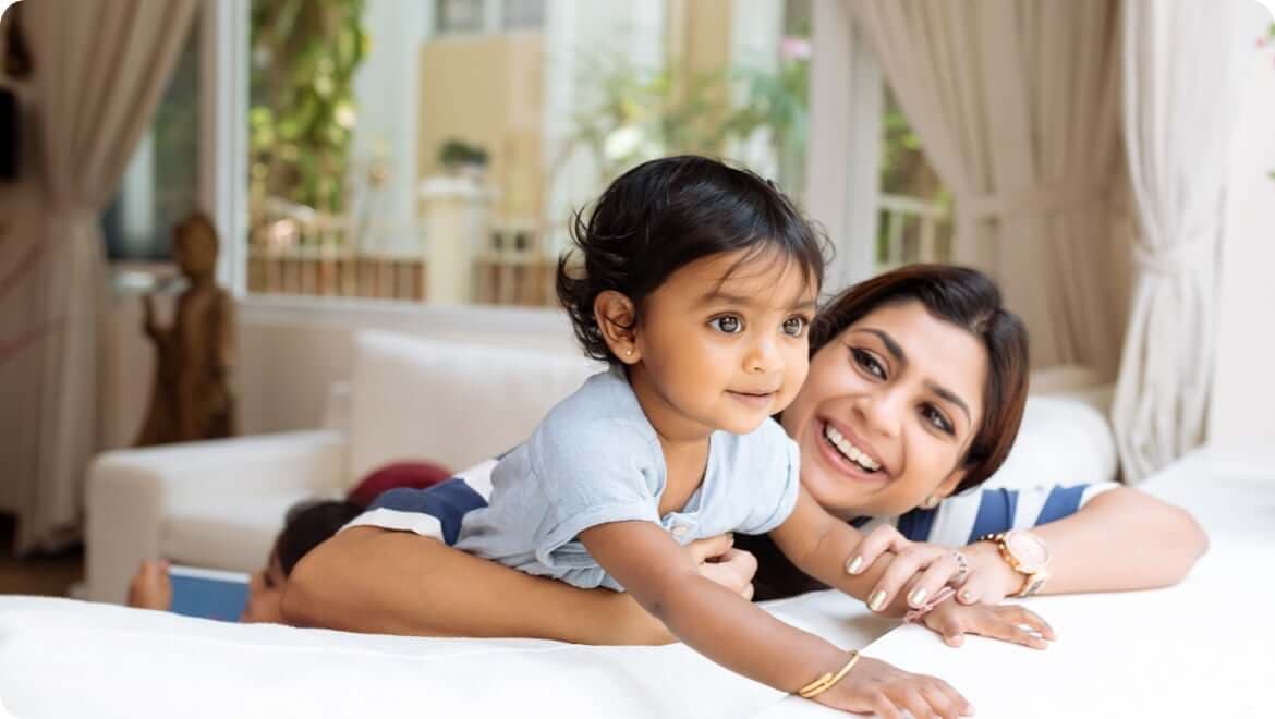A smiling woman lies on a couch supporting a baby who reaches forward on the sofa in a bright living room.