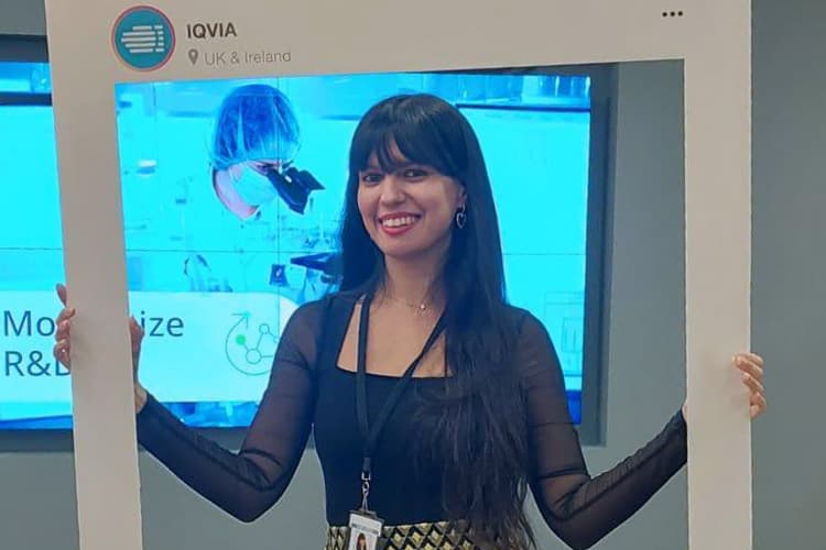 Woman smiling and posing with a photo frame prop at an IQVIA event, with a lab scene shown on a screen behind her.